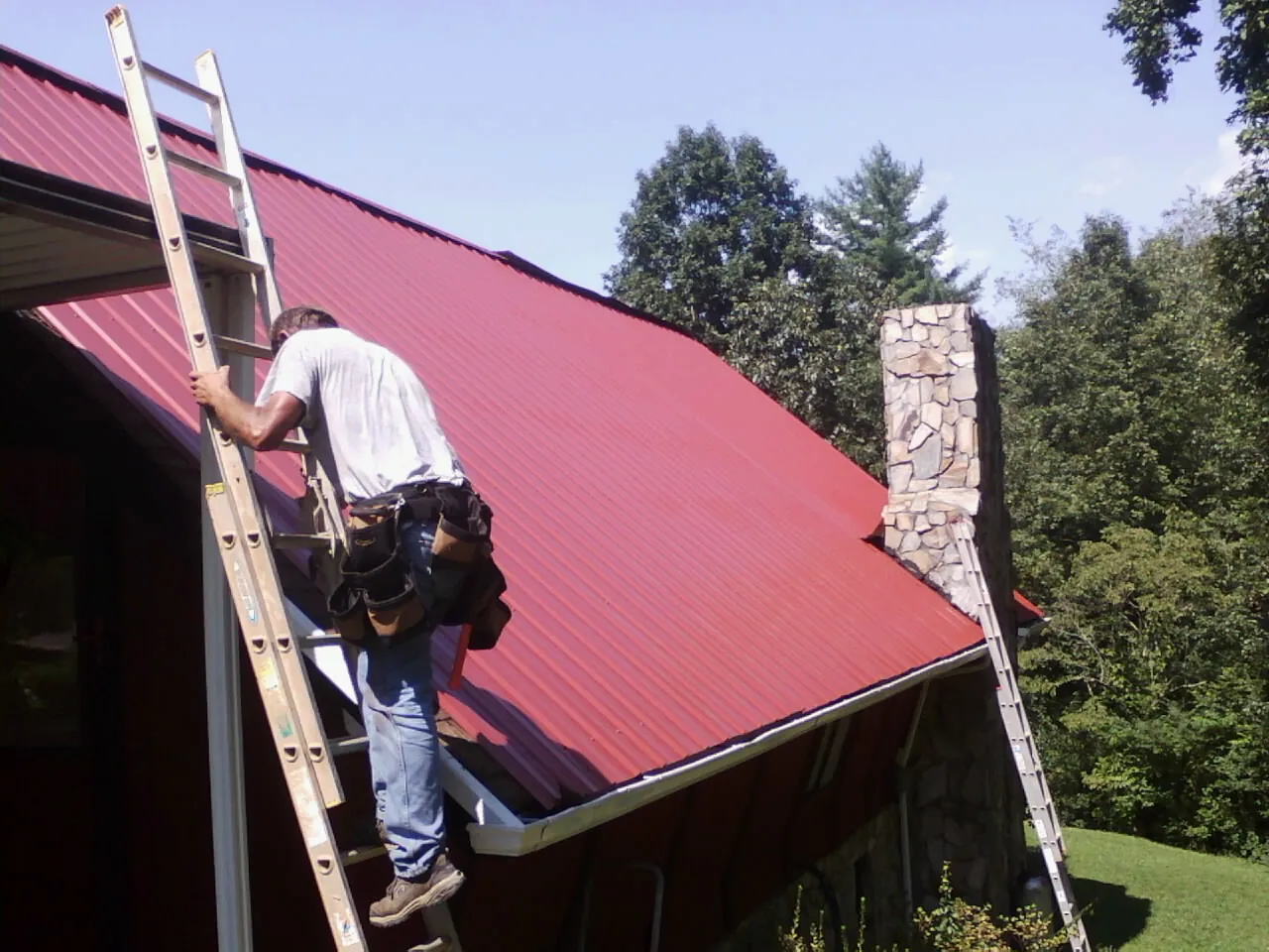 Roofer on a ladder beside a red metal roof and stone chimney — Soffit & Fascia Repair by Roofing Brothers in Carlos
